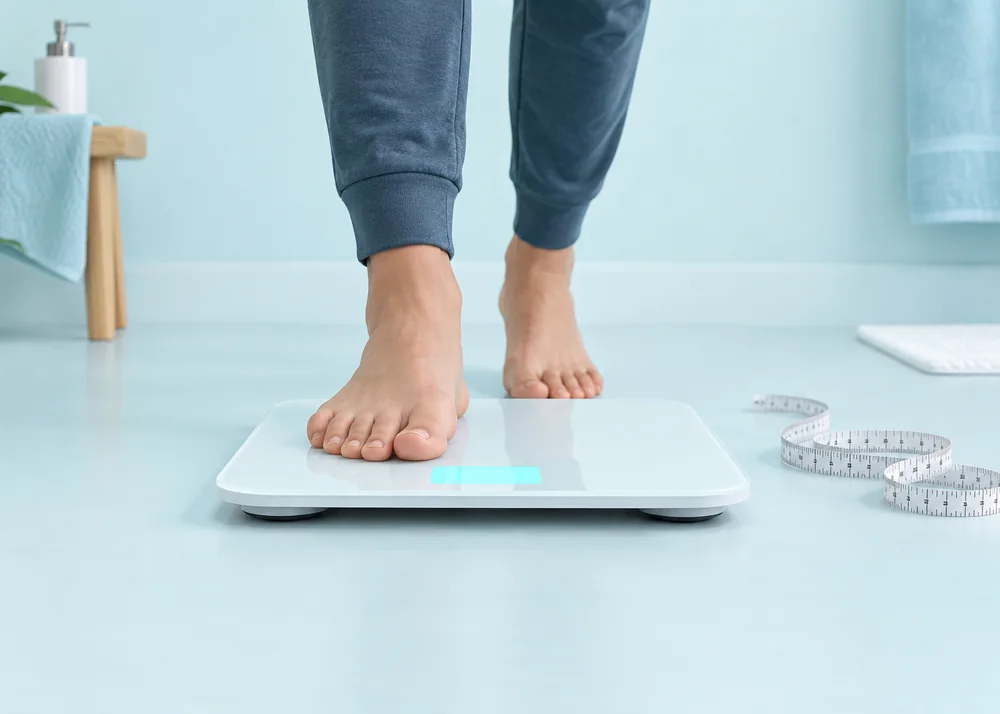 Bare feet stepping onto a white digital bathroom scale beside a measuring tape